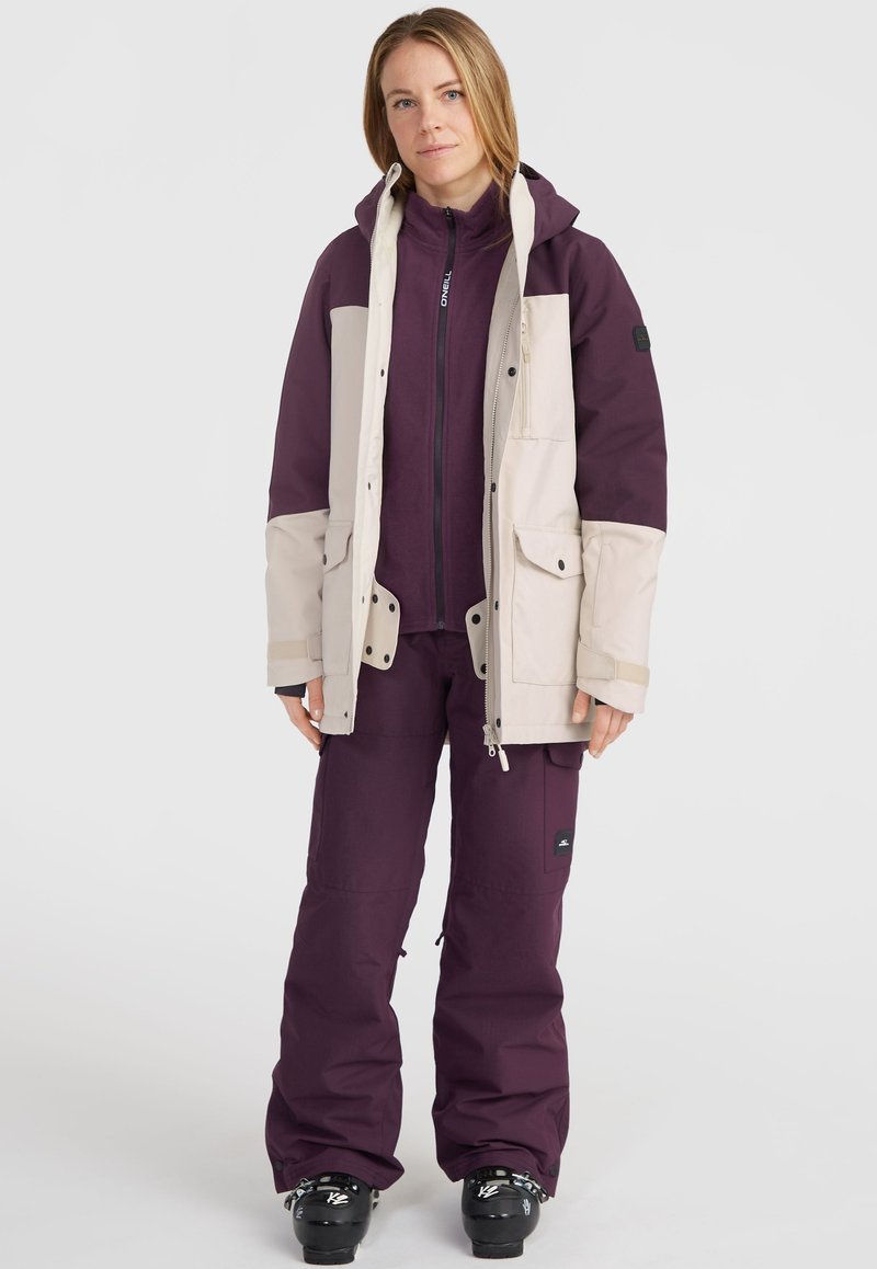 Skijacke und Hose Set in Lila und Creme. Die Jacke hat Taschen, eine Kapuze und Reißverschlüsse. Die Hose hat ein Logo-Emblem und eine lockere Passform. Strukturstoff.