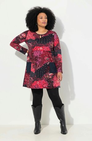 Robe à motif rouge et marine avec des manches longues, décolleté à ouverture clé et deux poches marines ; associée à des bottes de cowboy noires jusqu'aux genoux.