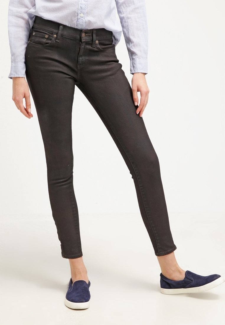 Persona che indossa jeans neri aderenti, una camicia a righe azzurra chiara e scarpe slip-on blu notte, in piedi con le gambe incrociate su uno sfondo bianco.