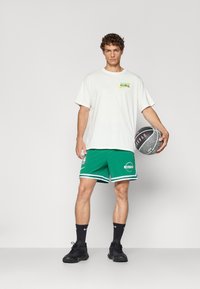 Hvid Nike t-shirt med et multicolor logo, grønne NBA Celtics shorts med logo, sorte basketballsko og holder en basketball.