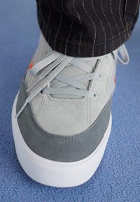 Sapatilha em camurça cinza e malha com sola branca, apresentando detalhes subtis em laranja, atacadores planos e um design de bico arredondado.