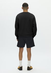 Sweat-shirt noir, coupe ample, col rond, associé à un short noir. Le modèle porte des chaussettes blanches et des baskets jaunes et noires. Vue de dos sur fond blanc.