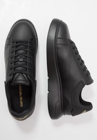 Black leather sneakers with a rounded toe, textured surface, flat rubber sole, and lace-up design. Gold branding inside the shoe.