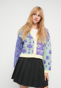 Γυναίκα με μακριά ξανθά μαλλιά, φοράει ένα κοντό ζακέτα με κουμπιά με μοτίβα σε μωβ, πράσινο και κίτρινο πάνω από ένα λευκό τοπ και μαύρη πλισέ φούστα.