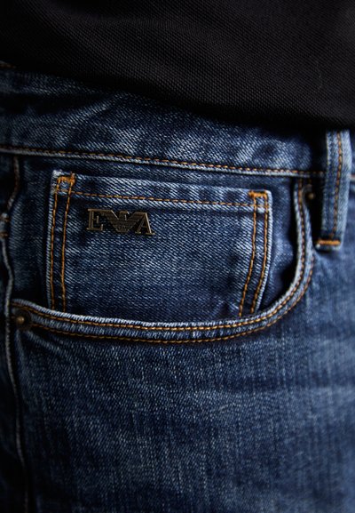 Jeans i mörkblått denim med synlig sömnad, med en liten svart logotyp på bakfickan och en klassisk femficksdesign.