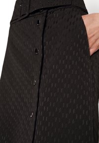 Detailní záběr na černou texturovanou sukni s předními cvoky a rukou zasunutou do postranní kapsy.