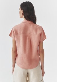 Short-sleeve, light pink linen shirt with a relaxed fit, rounded hem, and back pleat detailing. Features a classic collar and smooth texture.
