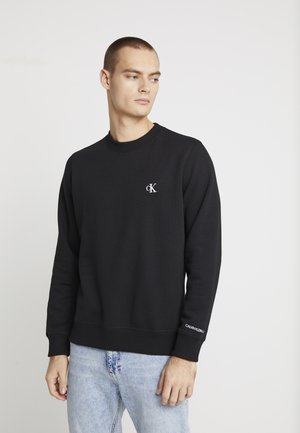 Bărbat purtând un sweatshirt negru Calvin Klein cu logo pe piept și pe mânecă, asortat cu blugi albastru deschis, privind în lateral.