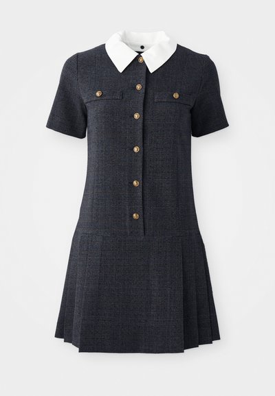 Rochie în carouri navy cu mâneci scurte, guler alb și cinci nasturi aurii. Designul fustei pliate adaugă textură și contrast.