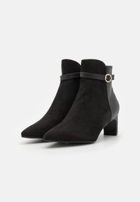 Botas de tobillo negras hechas de ante y cuero, con punta afilada, tacón medio y una correa decorativa con un acento metálico circular.