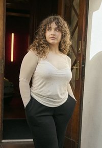 Beiges langärmliges Oberteil mit dezentem Glitzereffekt und entspanntem Schnitt, kombiniert mit schwarzen Hosen. Im Hintergrund ist eine Holztür und sanfte Beleuchtung zu sehen.