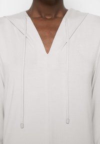 Personne à la peau foncée portant un sweat à capuche gris clair avec un col en V et des cordons lâches visibles au niveau de la poitrine.