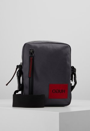 Petit sac bandoulière gris compact avec fermeture éclair noire, sangle réglable et patch rectangulaire rouge avec logo HUGO inversé sur le devant.