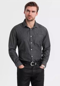 Chemise boutonnée gris foncé avec une poche, associée à un pantalon noir et une ceinture décorative. Le matériau semble texturé et décontracté.