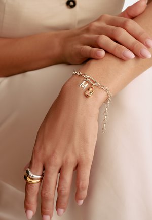 Hand einer Frau, die auf einem Arm ruht, trägt ein silbernes Gliederarmband mit dem Buchstaben "M" und einem kleinen Anhänger sowie einen zweifarbigen Silber-Gold-Ring am Finger.