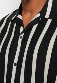 Gros plan d'une personne portant une chemise noire à rayures blanches verticales et boutons noirs, montrant le col et la partie supérieure de la poitrine.