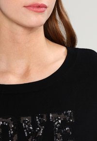 Schwarzer Pullover mit weitem Ausschnitt, der ein strukturiertes "LOVE"-Design aus schwarzen Pailletten aufweist. Glattes Strickmaterial mit figurnaher Silhouette.