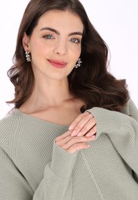 Világoszöld, bordázott kötött pulóver széles nyakkivágással. A modell csillogó fülbevalót visel, hosszú hullámos haja van, és ápolt körmei.