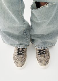 Sneakers feature a textured snakeskin pattern in shades of brown and tan, with black laces and a white rubber sole; paired with light blue jeans.