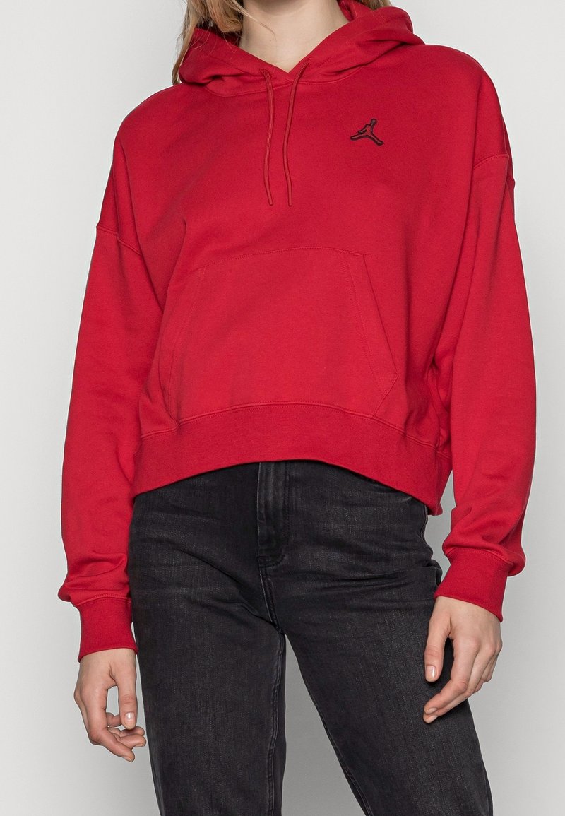 Personne portant un sweat à capuche court rouge avec cordons de serrage et un petit logo noir sur la poitrine, associé à un jean noir taille haute.