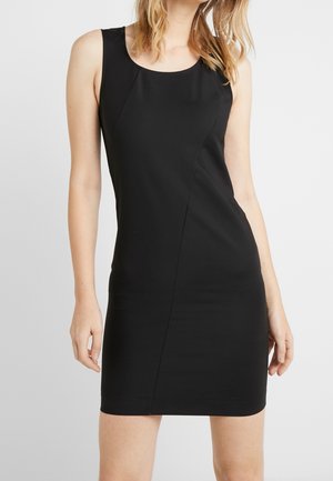 Vestido negro ajustado sin mangas con escote redondo, llevado por una mujer de pie frente a un fondo liso y claro.