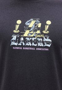 Μαύρο μπλουζάκι με vintage λογότυπο των LA Lakers, δύο τρόπαια πρωταθλήματος και το κείμενο "National Basketball Association" σε μωβ από κάτω.