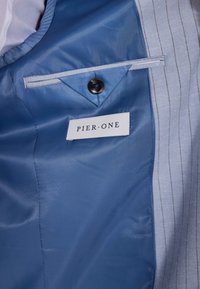 Das Innenfutter der blauen Jacke weist ein diagonal gestreiftes Muster auf, einen einzelnen schwarzen Knopf und ein weißes Etikett mit der Aufschrift "PIER•ONE". Sichtbare Kontrastnähte.