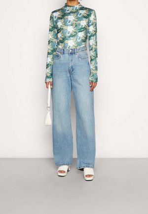 Langarm bloemen top in groen, blauw, crème, gecombineerd met high-waisted, lichtgewassen wijde jeans. Geaccessoriseerd met een witte handtas en hakken.