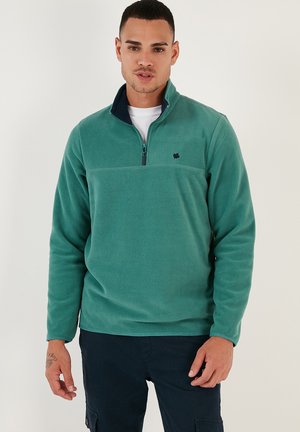Sudadera de forro polar verde con cuello alto y cremallera, que presenta un acento en color marino en el cuello y un pequeño logotipo en el pecho. Textura suave.