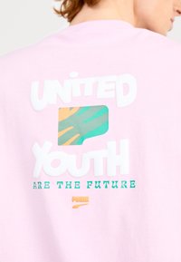 T-shirt di cotone rosa chiaro con testo audace in bianco e verde. Include un quadrato grafico multicolore con design astratto. Logo Puma in basso.