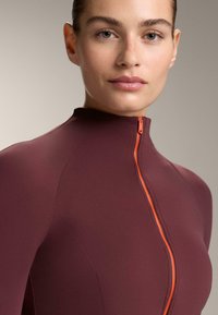 Bordó testhez simuló felső magas gallérral, kontrasztos narancssárga cipzárral. Simább textúra és varrás nélküli kialakítás, szabott illeszkedéssel.