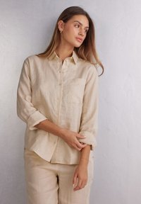 Camicia button-up in lino beige con taschino sul petto, maniche arrotolate e vestibilità rilassata. Il tessuto ha un aspetto morbido e leggermente strutturato.