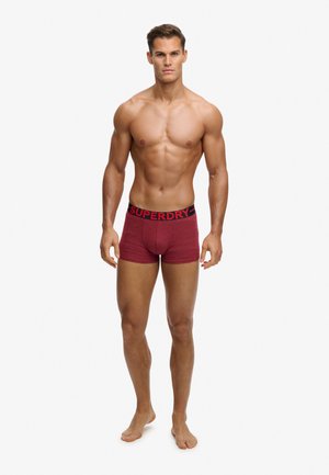 Gespierde man staat op blote voeten, draagt rode boxershorts met een zwarte tailleband waarop "Superdry" staat, tegen een effen witte achtergrond.