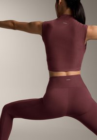 Top corto a coste bordeaux e leggings a vita alta. Tessuto liscio con una lieve lucentezza. Dettaglio logo vicino alla linea della vita. Design aderente.