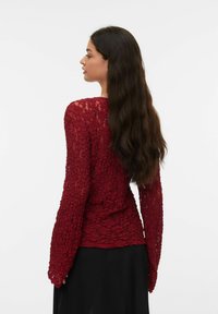 Czerwona koronkowa bluzka z długimi, rozkloszowanymi rękawami i fakturą w kwiatowy wzór. Widok od tyłu ukazuje dopasowaną sylwetkę, w połączeniu z czarną spódnicą.