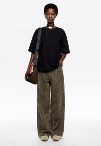 Maglietta nera oversize, pantaloni a gamba larga verde oliva con motivo, sneakers beige e una borsa a tracolla marrone con dettagli accentuati.