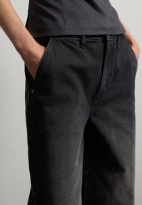Pantalones de mezclilla negra con un corte relajado, que cuentan con bolsillos frontales, un sutil desgaste y pasadores para cinturón que permiten una estilización ajustable.