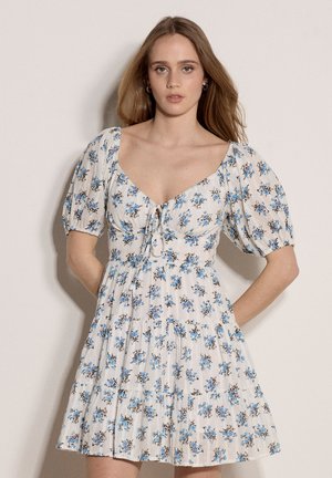 Junge Frau mit langen hellbraunen Haaren, die ein weißes Kleid mit blauem Blumenmuster und kurzen Puffärmeln trägt, steht vor einem schlichten Hintergrund.