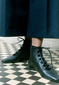 Juodi odiniai pusbačiai su smailiais pirštais, raišteliais ir plokščiu padu, dėvimi su tamsiai mėlyna sijonu ant languoto grindų.