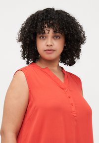 Blouse sans manches rouge avec un col à encoche, fermeture à boutons sur le devant et un tissu léger et texturé.