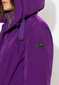 Giacca isolante viola con cappuccio, caratterizzata da una finitura testurizzata, zip frontale e un'etichetta nera con logo sul braccio.
