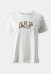 Vit bomull t-shirt med en "GAP"-logotyp i fet beige text, prydd med rosa blommönster under texten. Klassisk passform.
