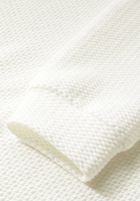 Weißer, strukturierter Stoff mit einem erhöhten Waffelmuster, der einen umgeschlagenen Bund aufweist. Das Material wirkt weich und leicht und ist für legere Kleidung geeignet.