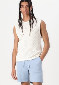 Pull en tricot sans manches de couleur crème avec un motif en diamant, associé à un short bleu clair avec des poches avant et une taille à cordon.