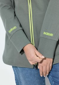 Chaqueta gris con cremallera y acentos amarillos en la cremallera y puños, con el texto "STARBOARD" y "BACKBOARD" en las mangas. Textura suave.