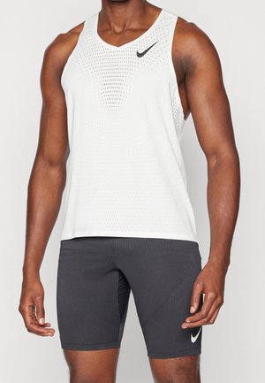 Homme portant un débardeur de sport blanc avec logo Nike noir et un short noir moulant avec logo Nike blanc sur fond uni.