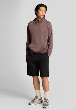 Sweatshirt à capuche mauve foncé avec une poche avant et un logo, associé à un short noir. Porté avec des chaussettes beiges et des baskets grises.