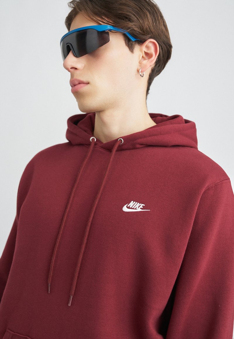 Bordo mikina s kapucňou na šnúrku, s malým bielym logom Nike. Model nosí čierne slnečné okuliare s modrými akcentmi.