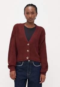 Burgunderfarget strikkegenser med dyp V-hals, som har tre dekorative knapper og ballongsleeves, kombinert med mørke denim jeans.