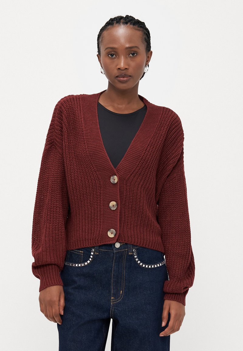 Bordeaux gebreide cardigan met een diepe V-hals, voorzien van drie decoratieve knopen en ballonmouwen, gecombineerd met donkere spijkerbroeken.